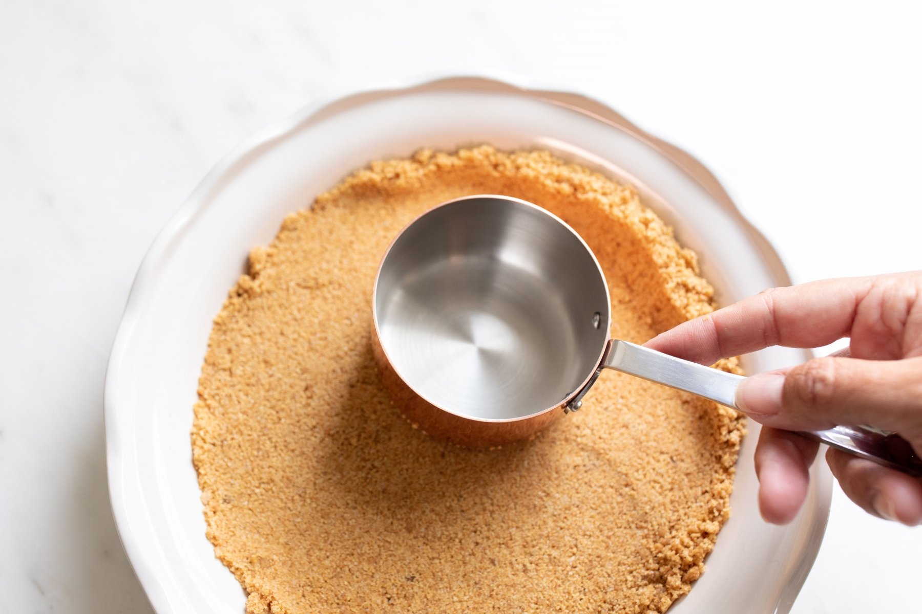 A hand with a copper measuring cup on a homemade graham cracker crust.
