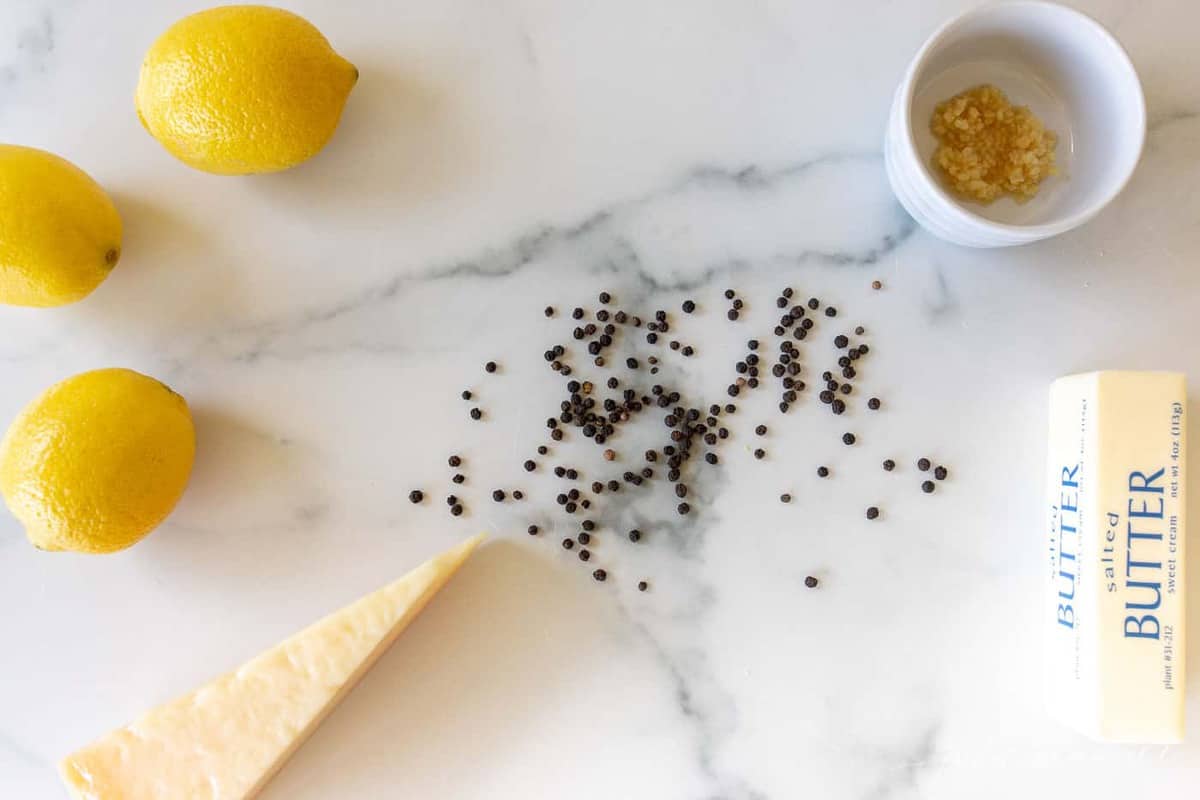 A marble surface with ingredients for lemon pepper sauce, including coarse black pepper, whole lemons, garlic, and cheese.