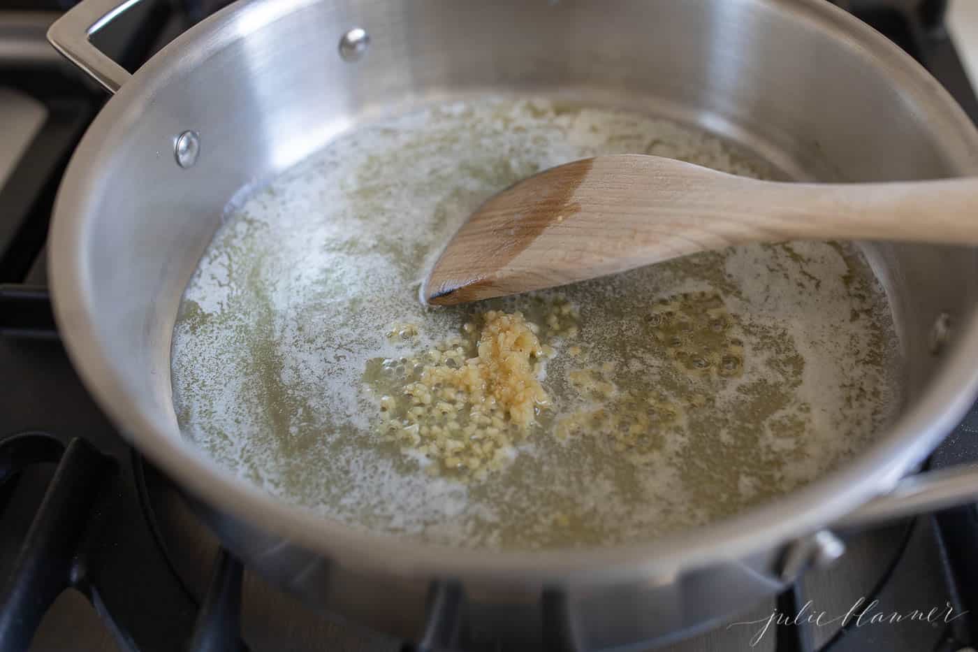 A silver pan on a stove top, melted butter and garlic being stirred with a wooden spatula.