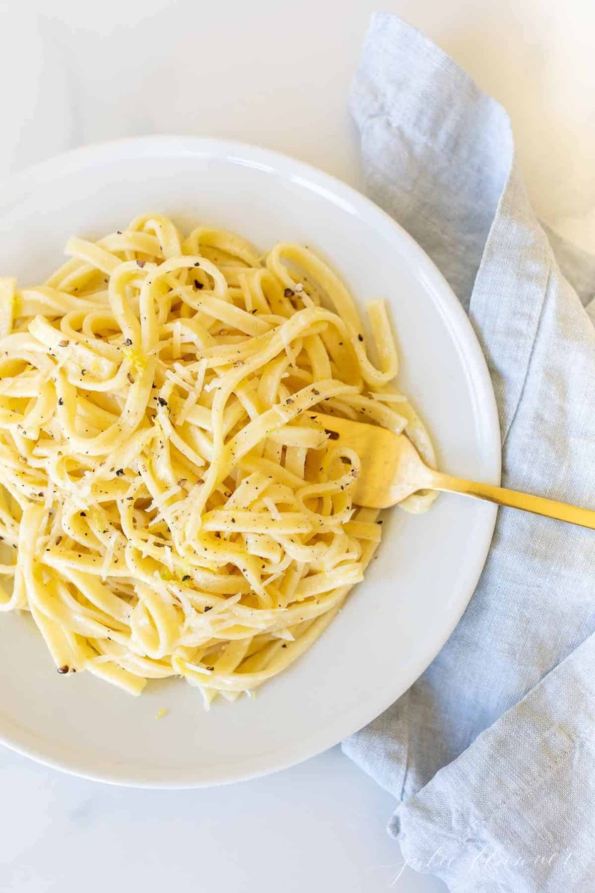 A white plate with a bowl full of lemon pepper pasta, gold fork to the side.