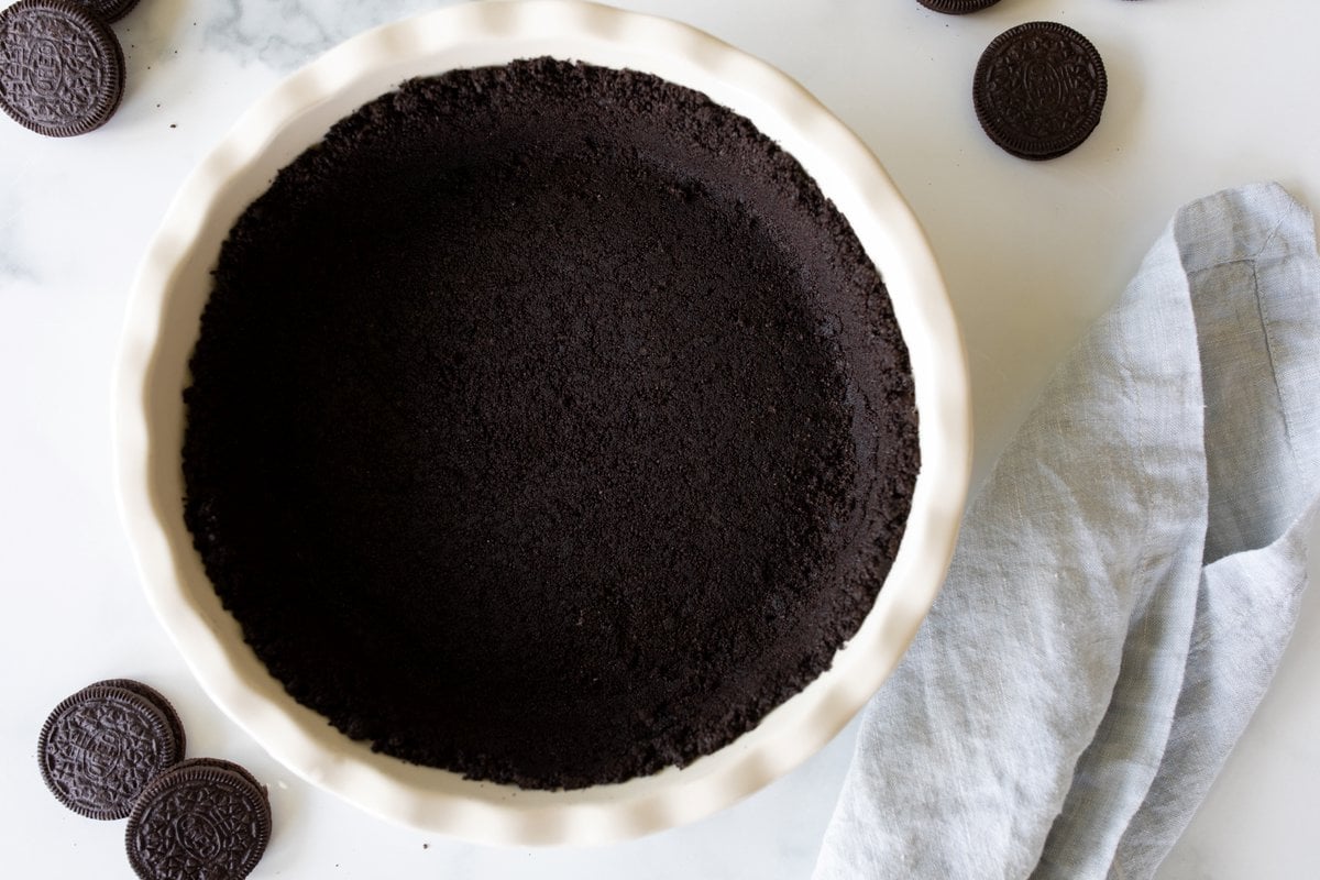 A white ceramic pie dish with an oreo crust recipe inside, oreos to the side.