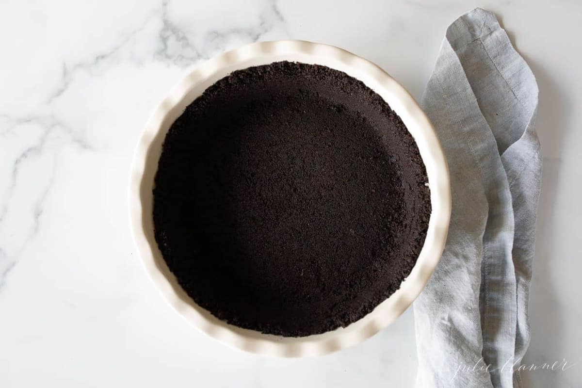 A white ceramic pie dish with an oreo cookie crust inside, blue linen towel to the side.