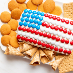 A red white and blue snack made of Funfetti dip in a flag shape, on a plate of cookies and crackers.