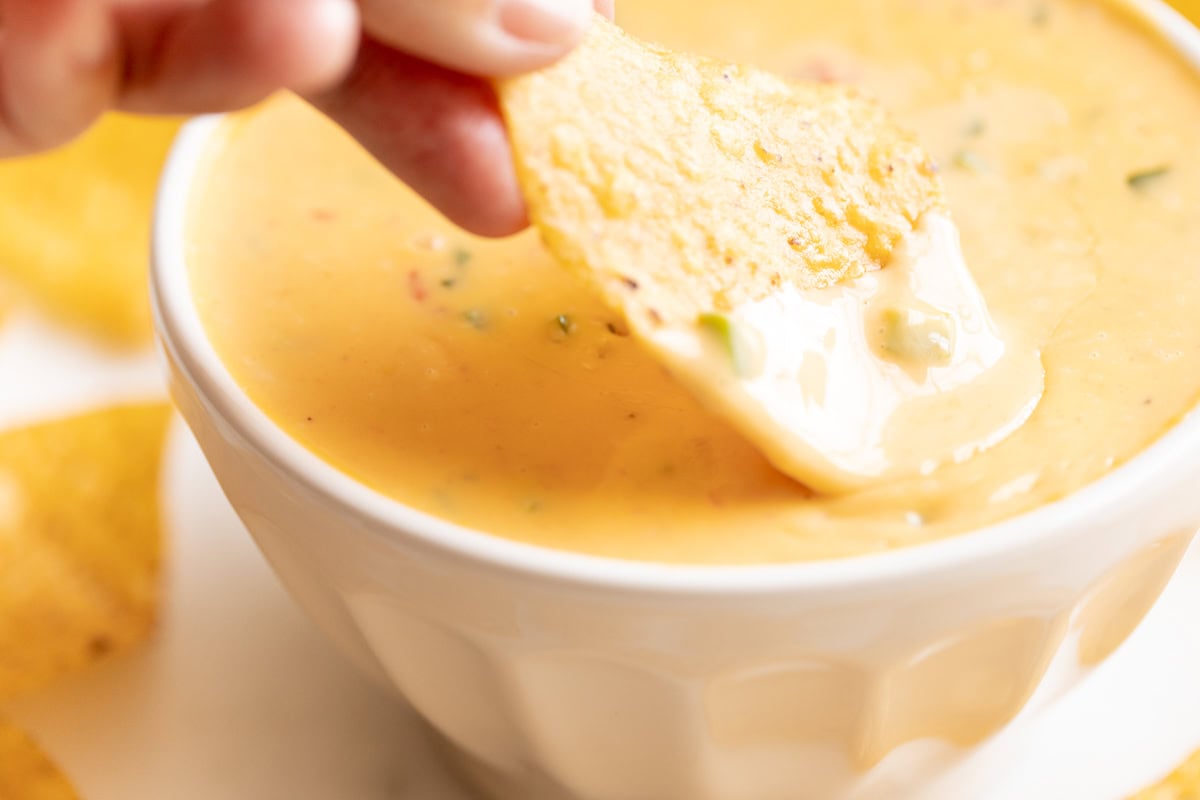 A hand dips a tortilla chip into a bowl of velveeta cheese dip, rich and creamy with visible herbs and spices.