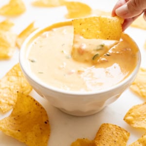 A hand dips a tortilla chip into a bowl of velveeta cheese dip, with more chips scattered around on a white surface.