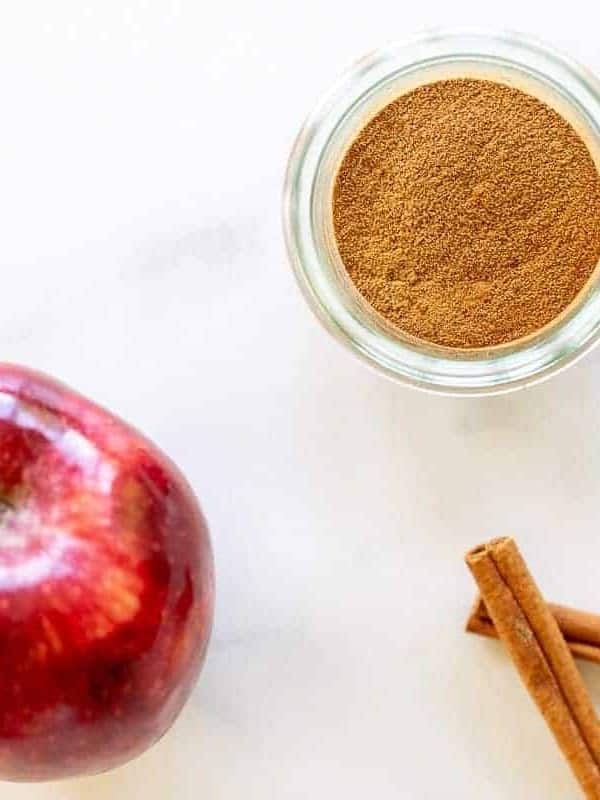 A marble surface with a small jar of apple pie spice, cinnamon sticks and an apple to the side.