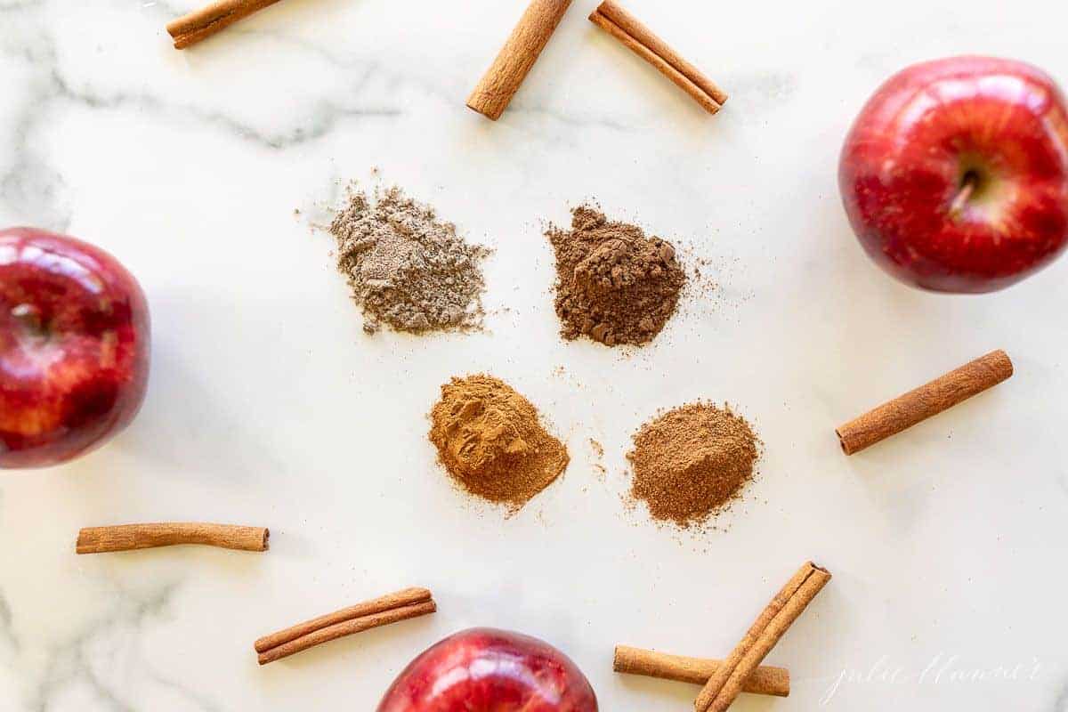 A marble surface filled with seasoning piles, cinnamon sticks and apples to make an apple pie spice recipe.