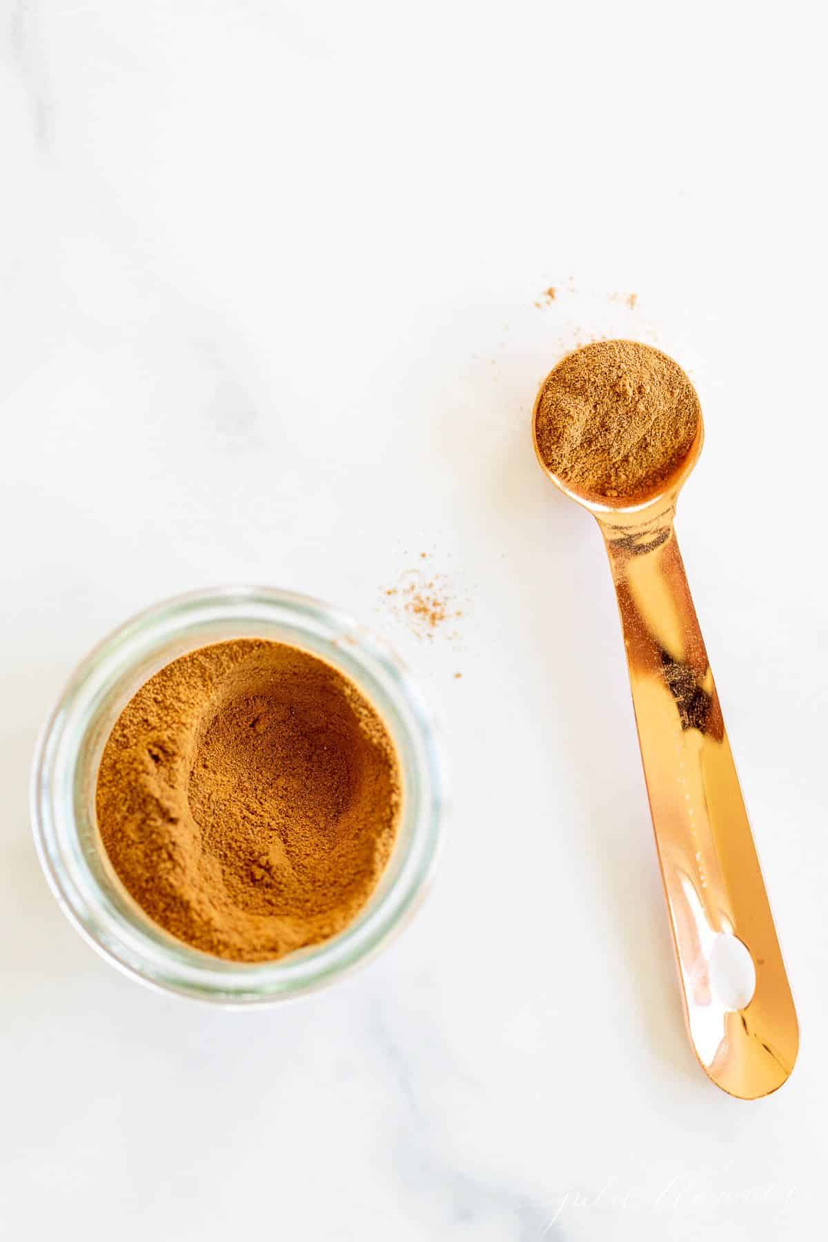 A marble surface with a clear jar full of apple pie spice and a teaspoon to the side.