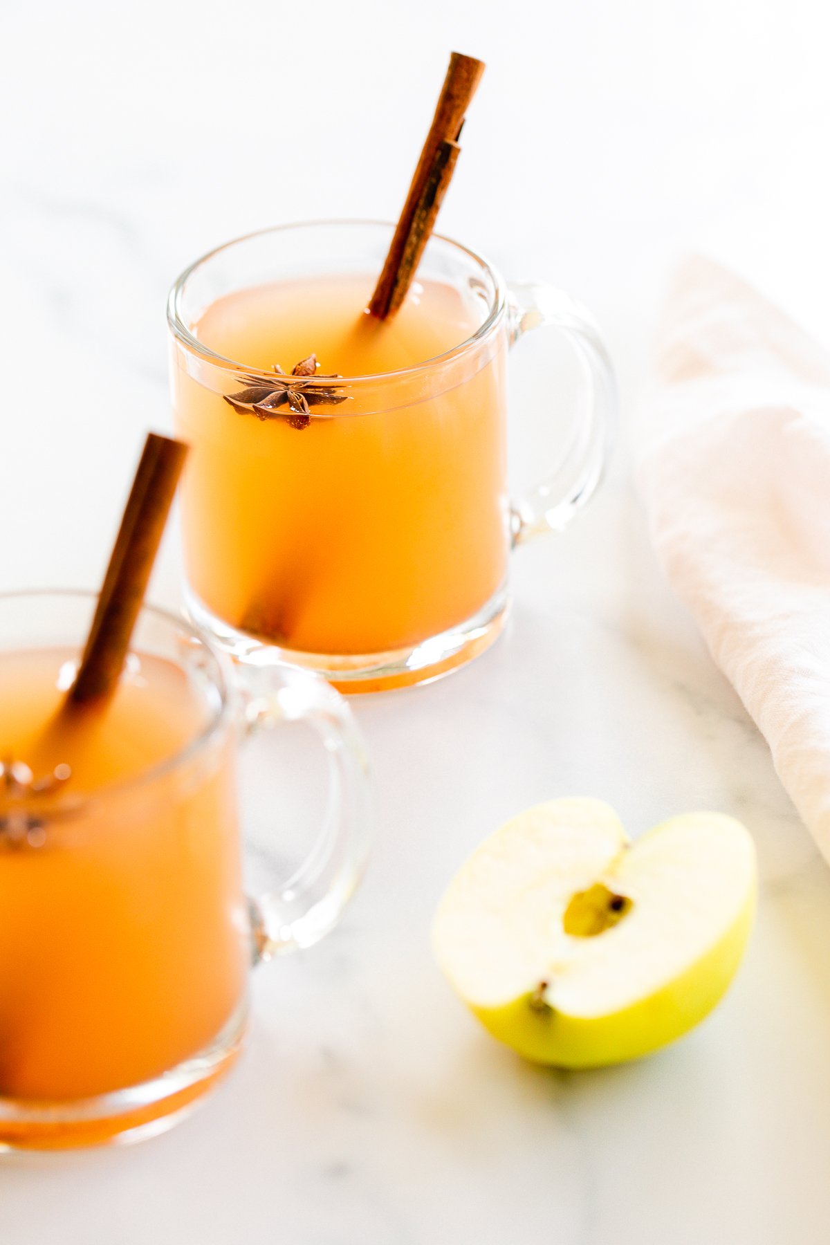 A clear glass mug full of homemade apple cider, with apples in the background.