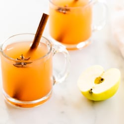 A clear glass mug full of homemade apple cider, with apples in the background.