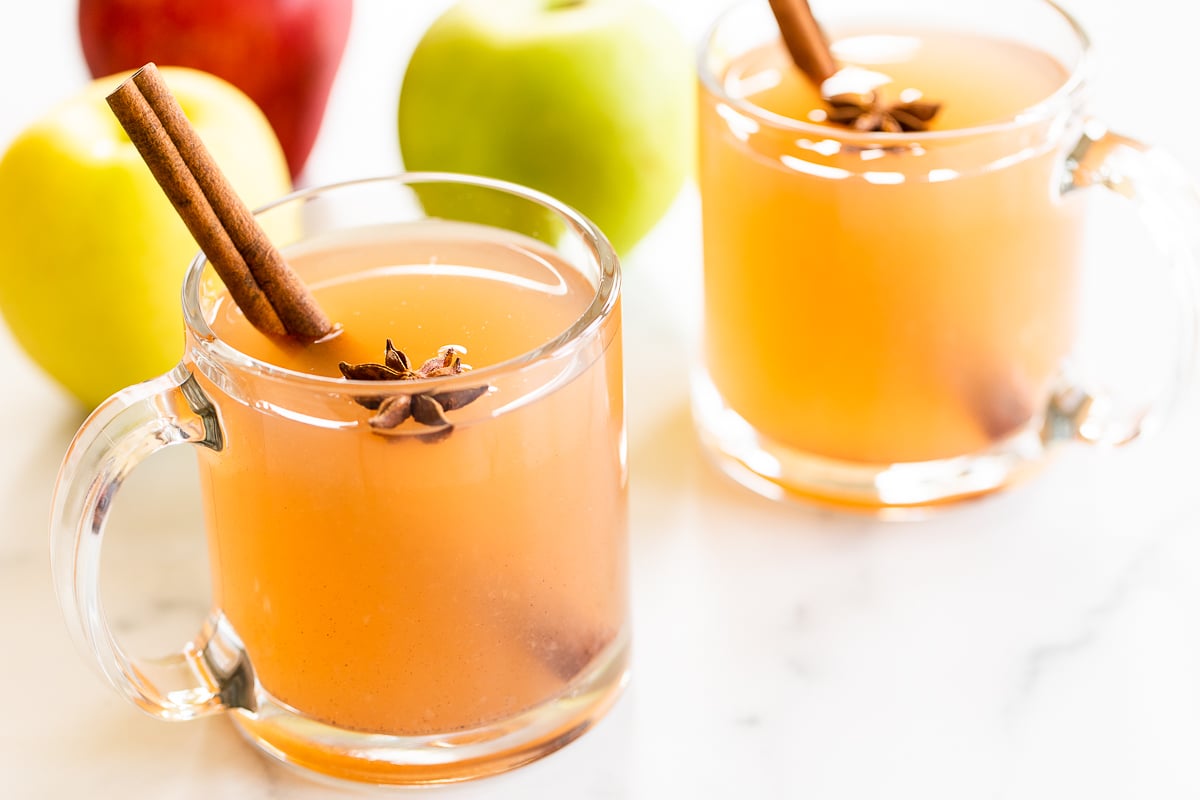 A clear glass mug full of homemade apple cider, with apples in the background.