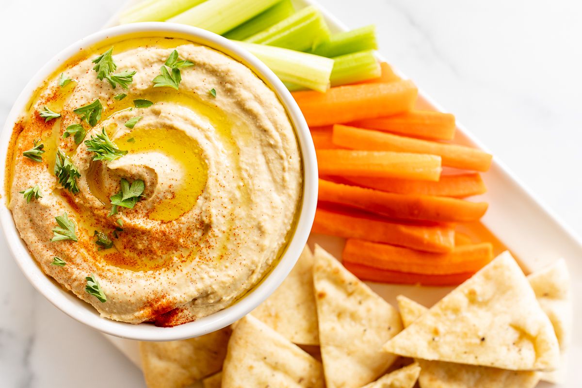 A white bowl of hummus surrounded by a platter of vegetables for dipping. 