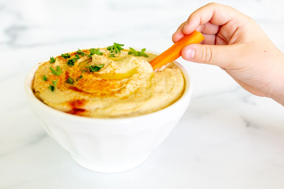 A small bowl of an easy hummus recipe on a white countertop, hand dipping a carrot inside.