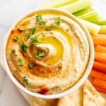 A white bowl of hummus surrounded by a platter of vegetables for dipping.
