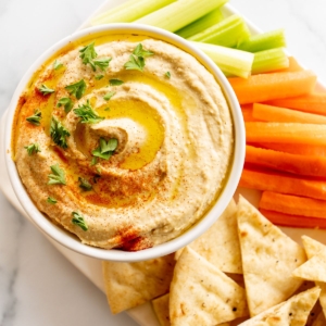 A white bowl of hummus surrounded by a platter of vegetables for dipping.