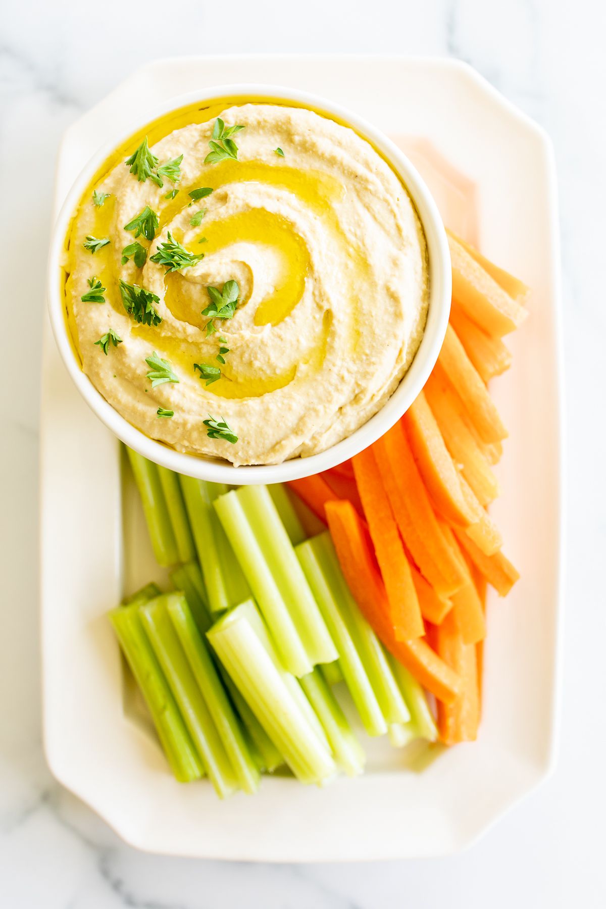 A white bowl of hummus surrounded by a platter of vegetables for dipping.