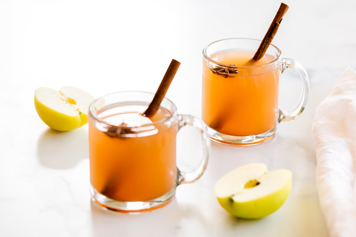 A clear glass mug full of homemade apple cider, with apples in the background.