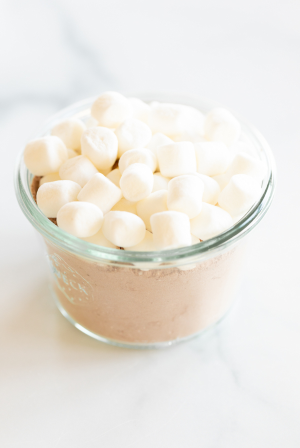 Homemade hot chocolate with marshmallows in a glass bowl on a white surface.