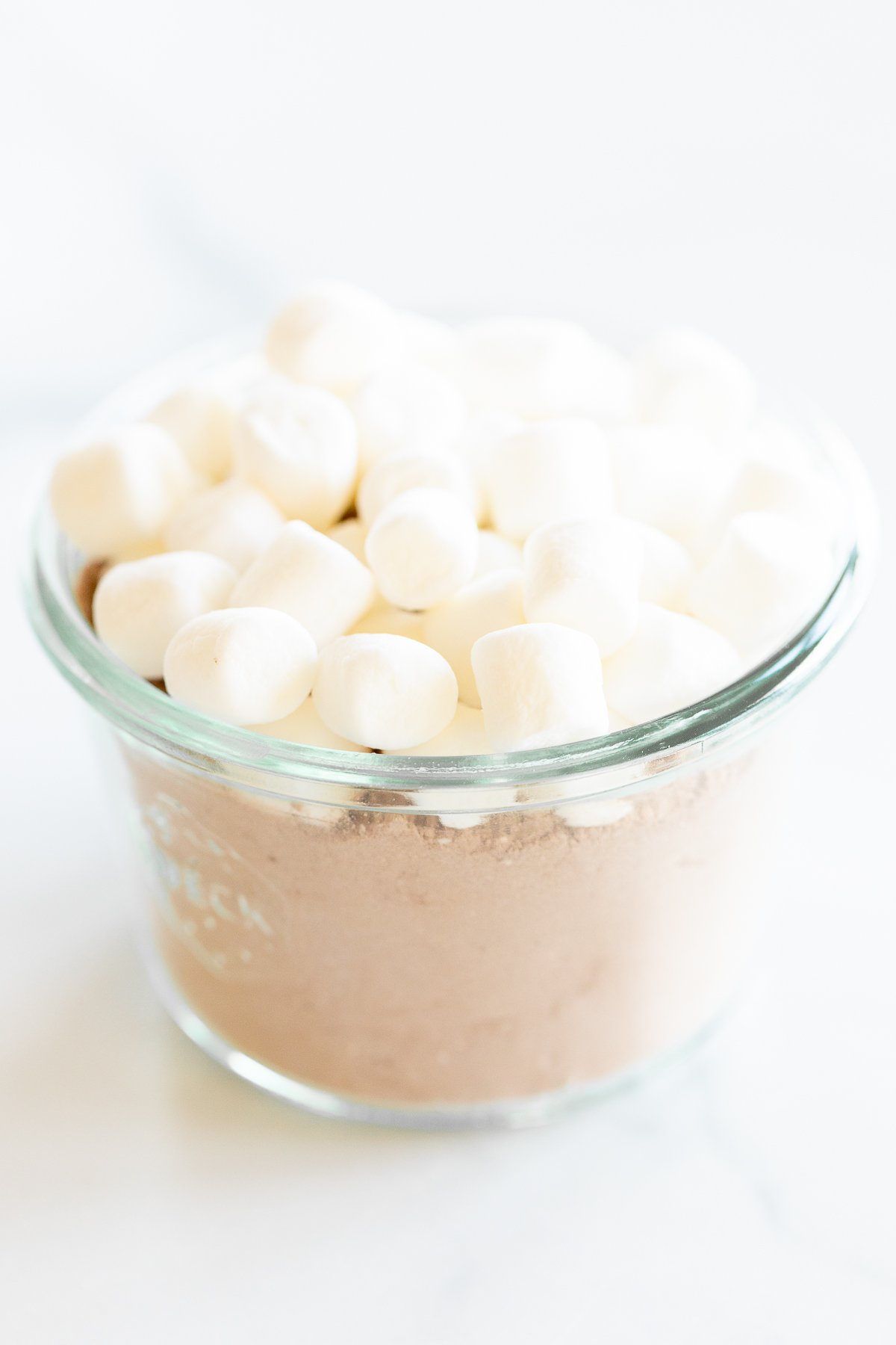 Homemade hot chocolate with marshmallows in a glass bowl on a white surface.