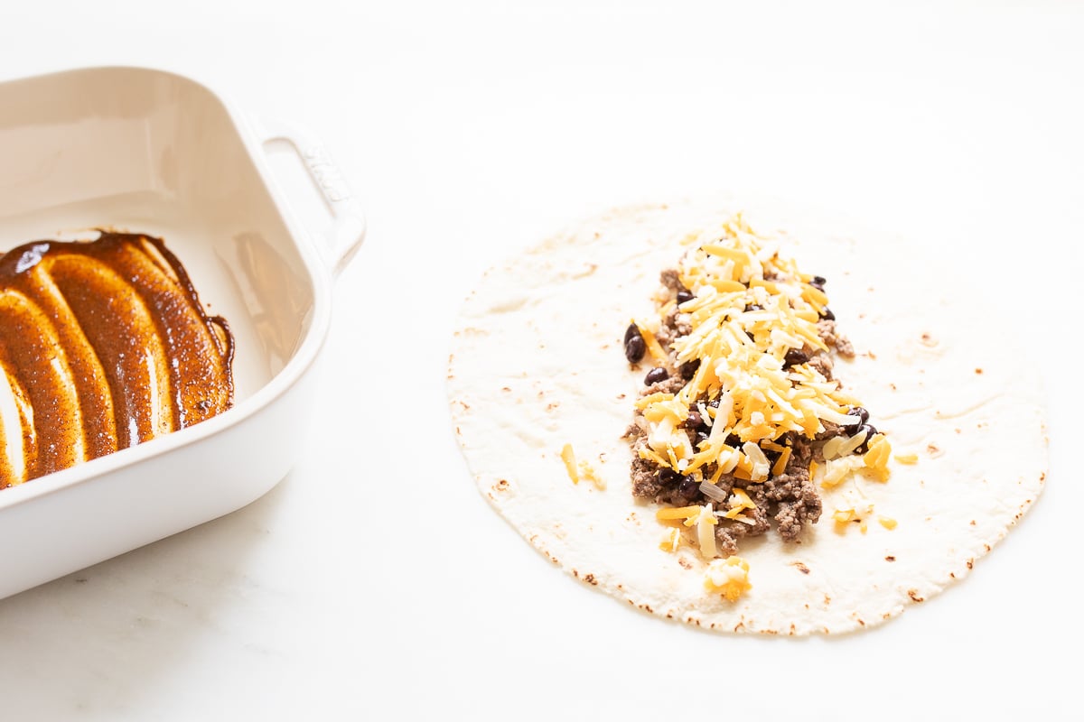 A tortilla topped with beef and cheese, next to a pan full of enchilada sauce