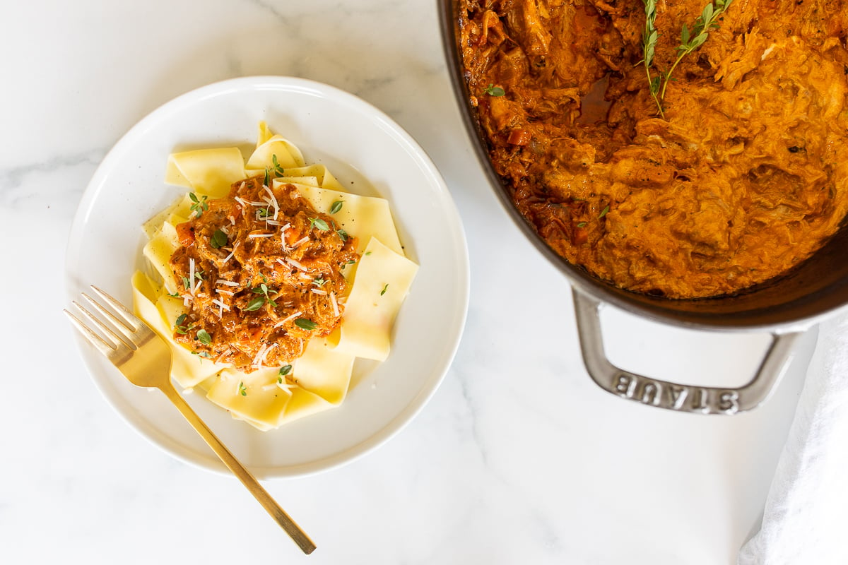 A plate of pork ragu served over pasta, with a cast iron skillet of sauce in the background