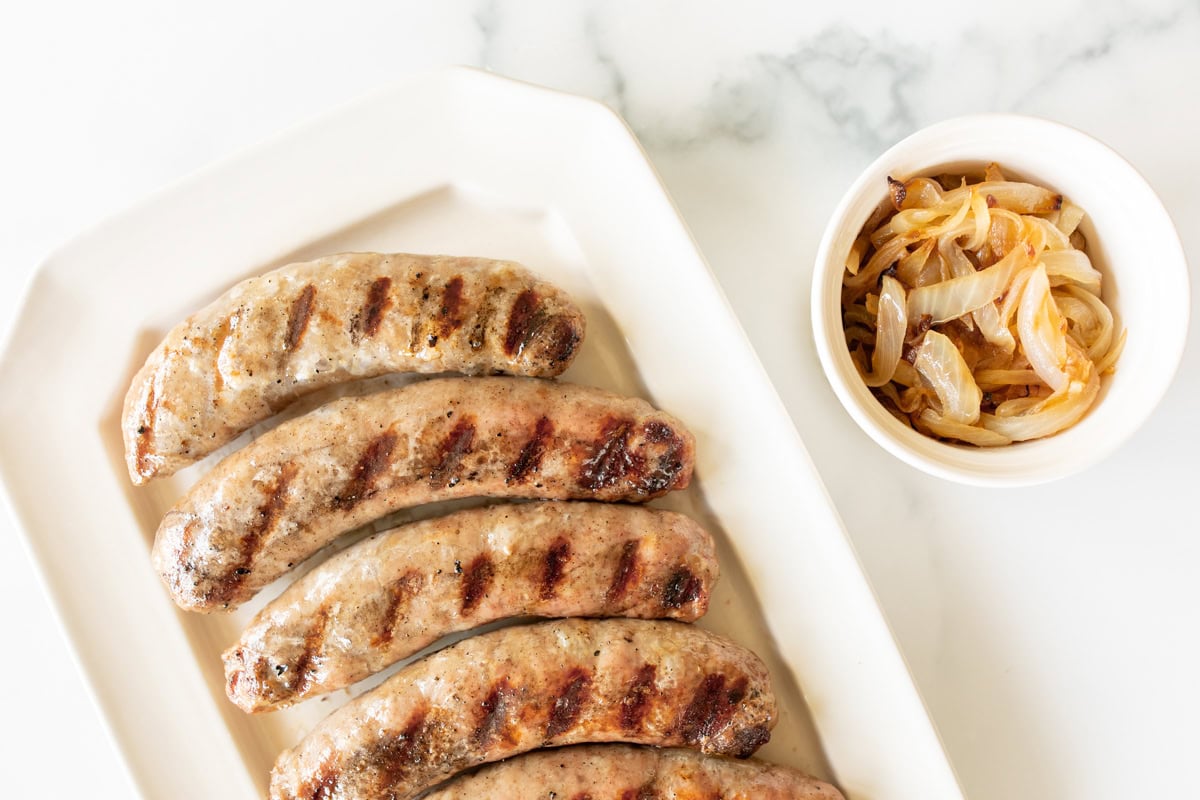 A white rectangular plate showcases five perfectly grilled bratwursts next to a small bowl of sautéed onions on a marble surface, exemplifying expert cooking techniques.