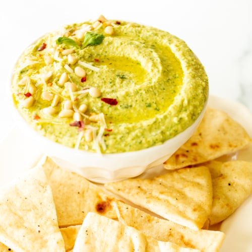 Pesto hummus in a white bowl surrounded by pita bread.