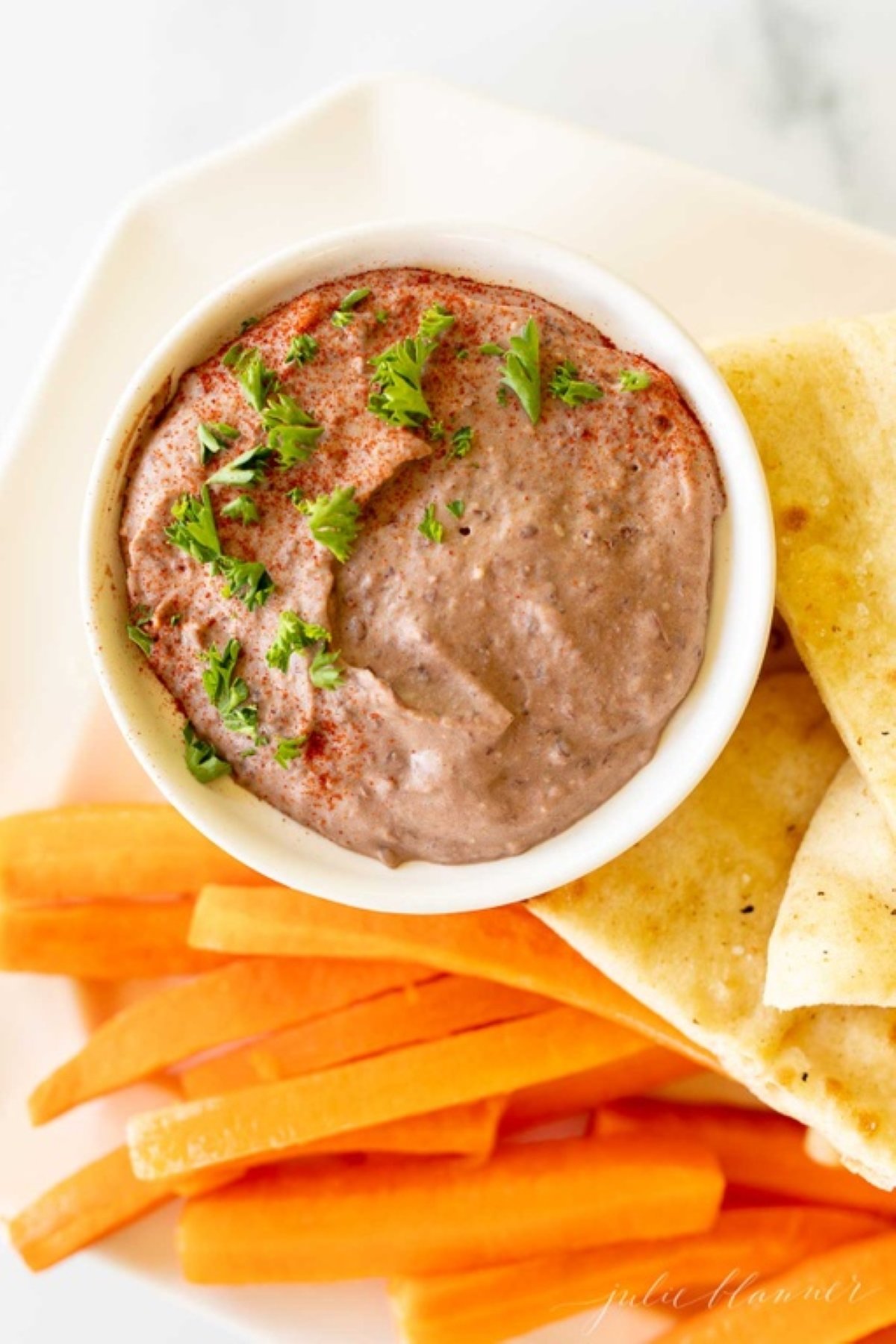 A white platter with pita slices, cut carrots and celery, with a small white bowl of black bean hummus.