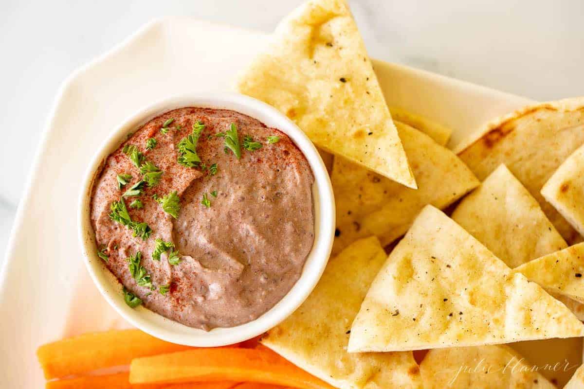 A white platter with a bowl of black bean hummus and pita triangles, celery and carrots.