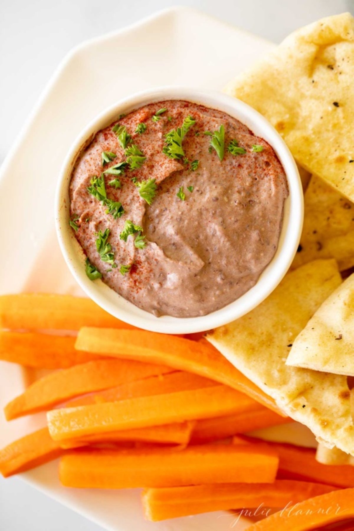 A white platter with pita slices, cut carrots and celery, with a small white bowl of black bean hummus.