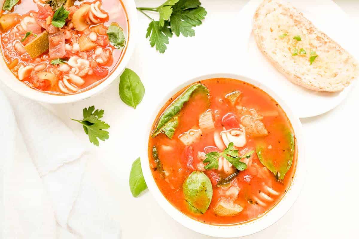 A white bowl of minestrone soup, another bowl in the background with herbs and a slice of bread on the surface.