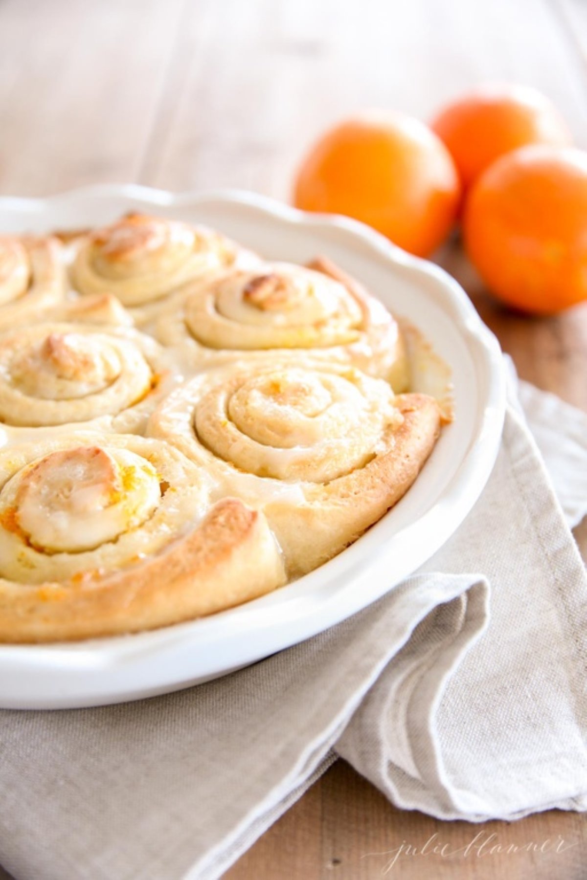 Orange rolls baked into a white ceramic round dish. Fresh oranges are on the table in the background.