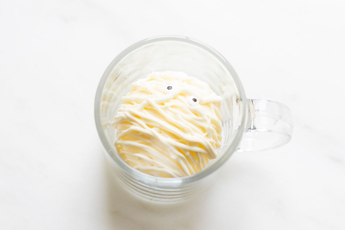 A white chocolate hot cocoa bomb decorated as a mummy, inside a clear glass mug