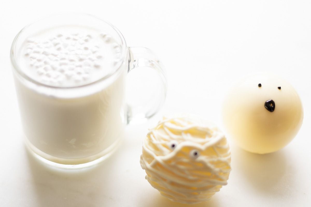 A white chocolate Halloween hot chocolate bomb, decorated as a mummy. Next to it is a glass mug full of white chocolate hot cocoa.