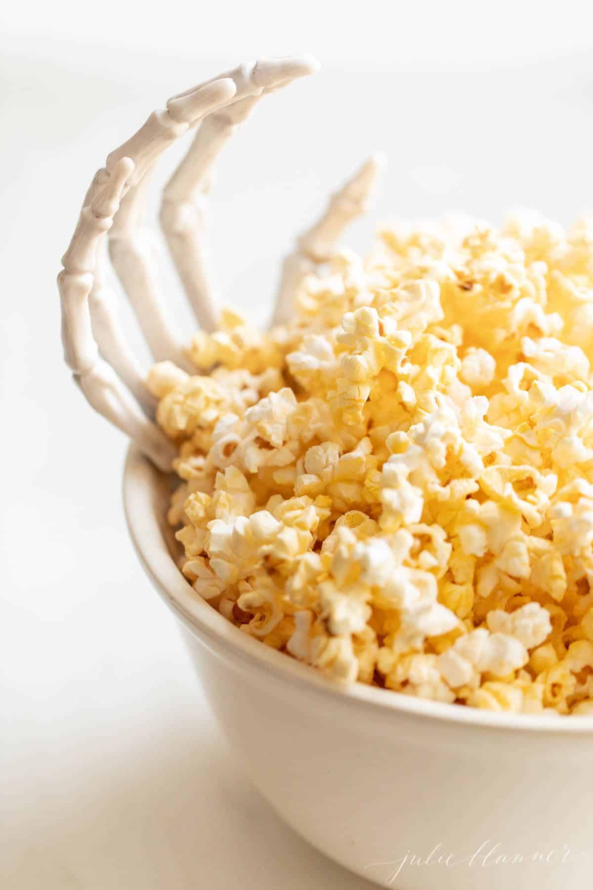 A white bowl full of popcorn with a skeleton hand reaching out for Halloween decorations.