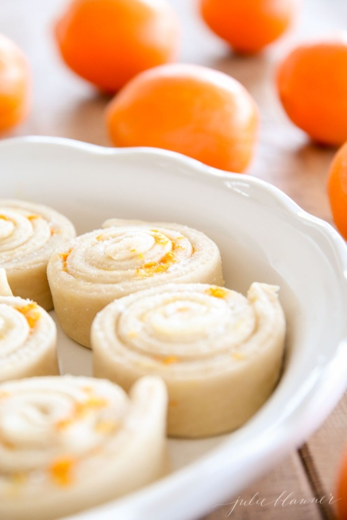 An orange rolls recipe, before baking, placed in a white ceramic round dish. Fresh oranges are on the table in the background.