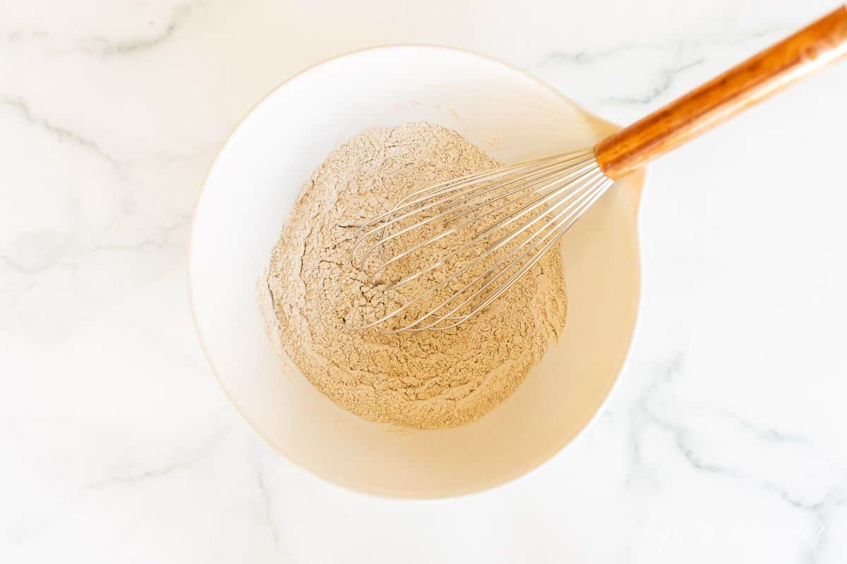 A white bowl full of ingredients for baking, whisk inside bowl.