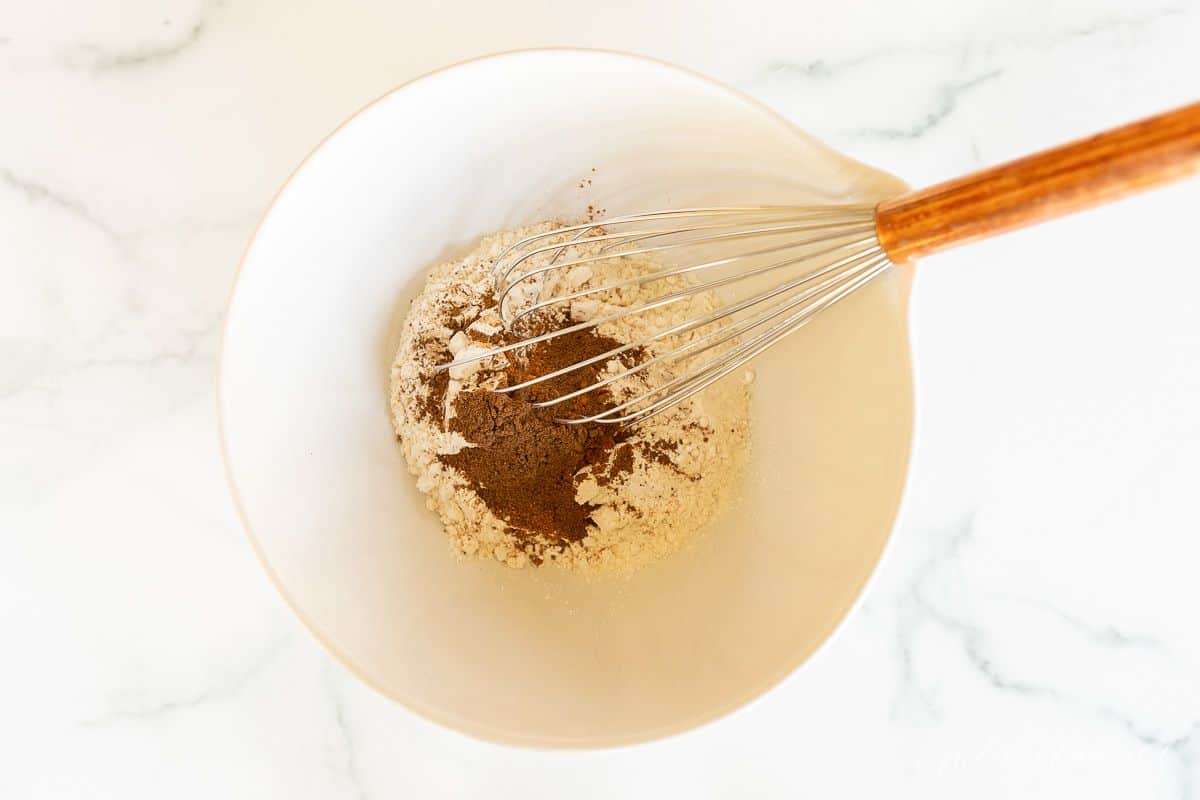 A white bowl full of ingredients for baking, whisk inside bowl.