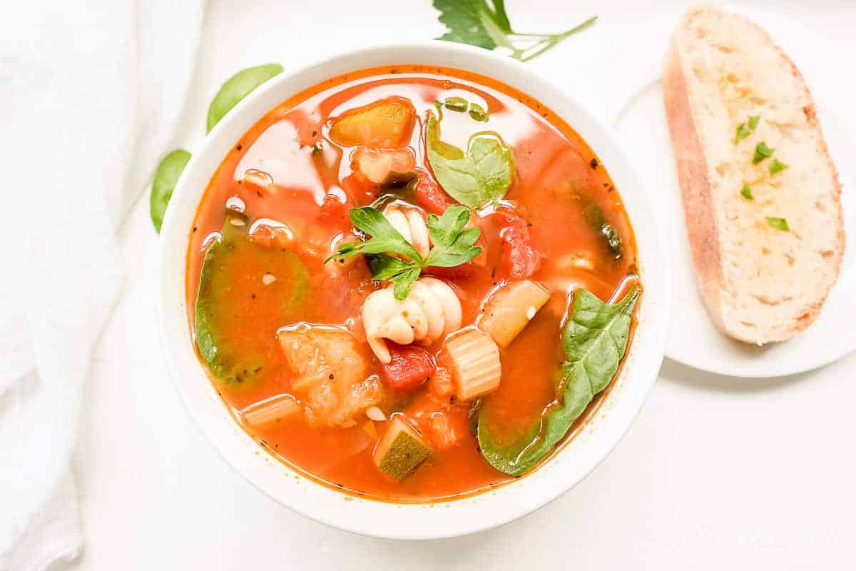 A white bowl of minestrone soup with herbs and a slice of bread on the surface.