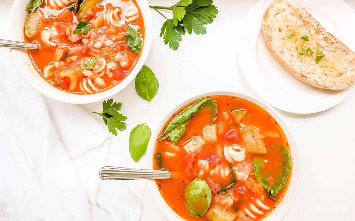 A white bowl of minestrone soup, another bowl in the background with herbs and a slice of bread on the surface.