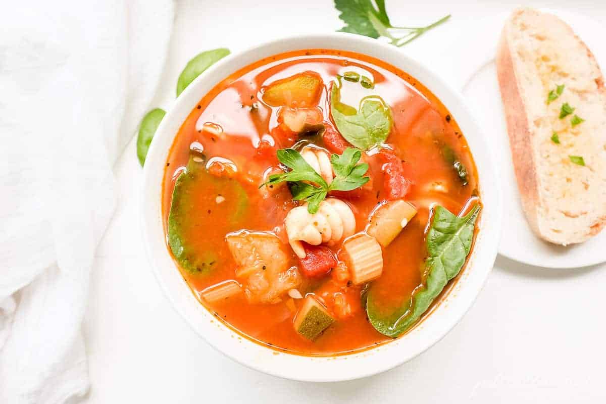 A white bowl of minestrone soup with herbs and a slice of bread on the surface.