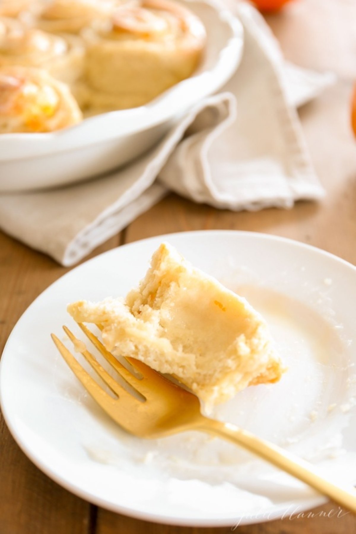 A small bite of an orange roll on a white plate, baking pan full of the orange roll recipe in the background.