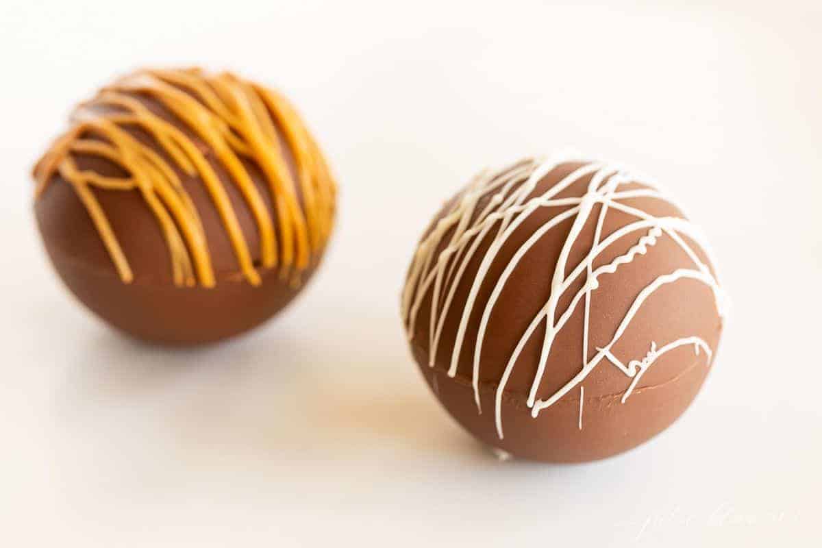 Two peanut butter hot chocolate bombs on a marble surface, drizzled with peanut butter and white chocolate.