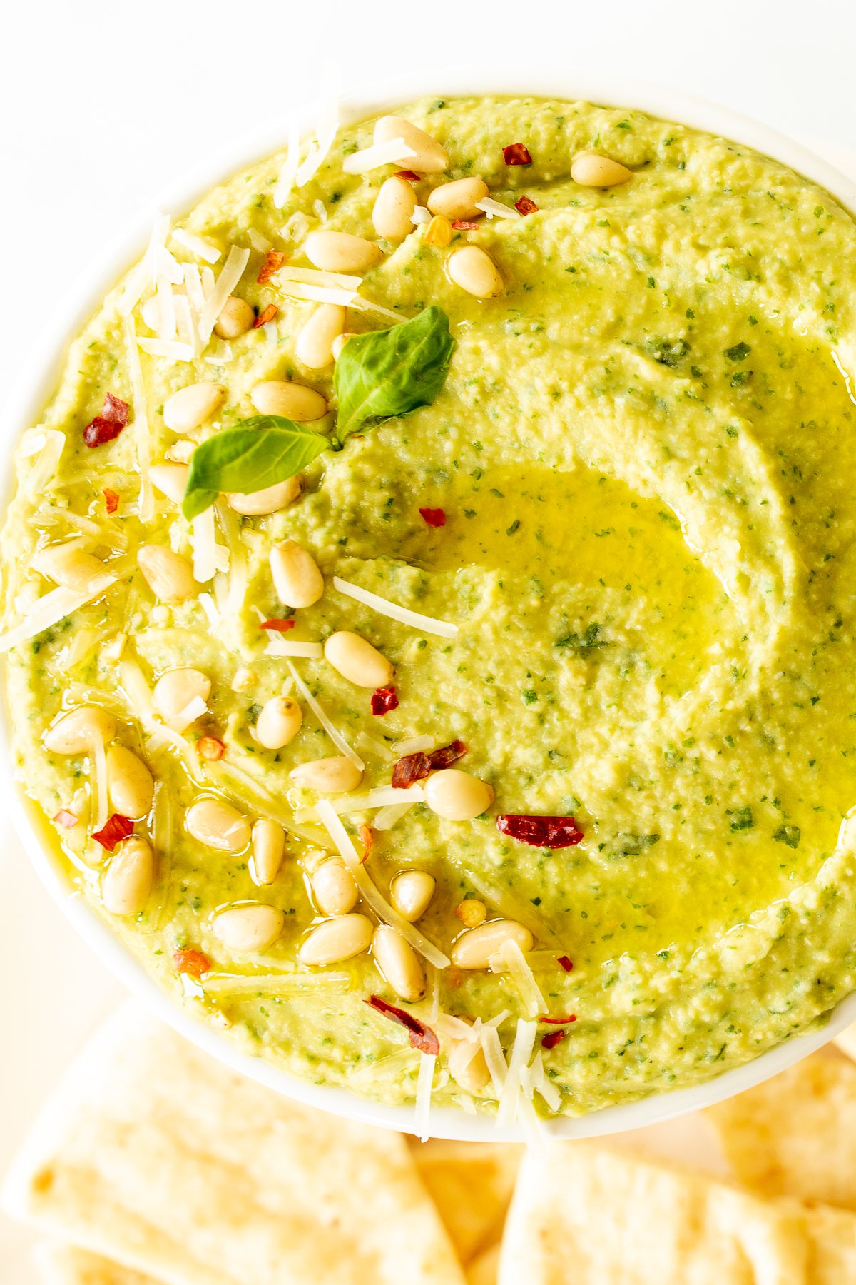 A white bowl full of basil pesto hummus, topped with pine nuts and parmesan.