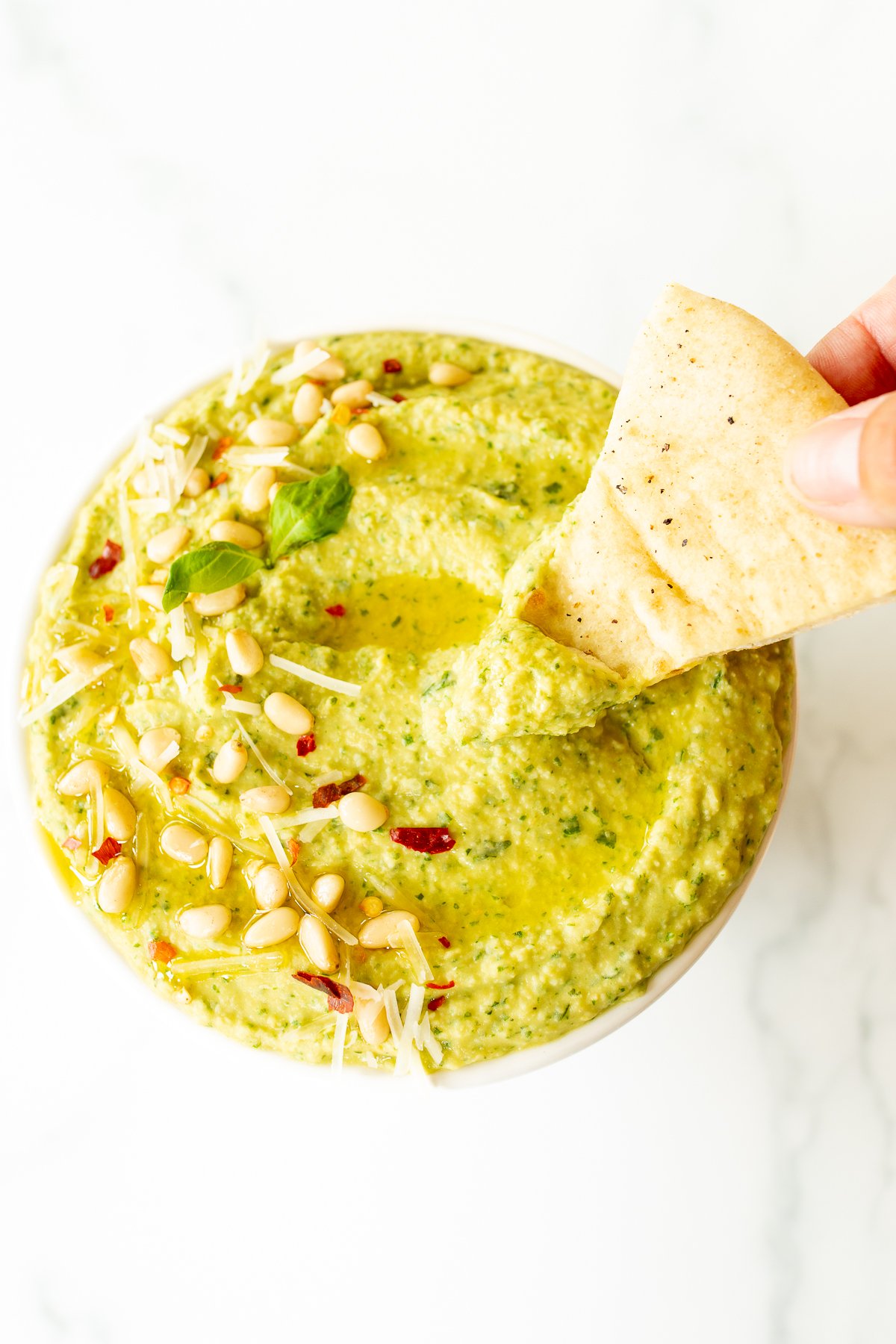 A white bowl full of basil pesto hummus, topped with pine nuts and parmesan.