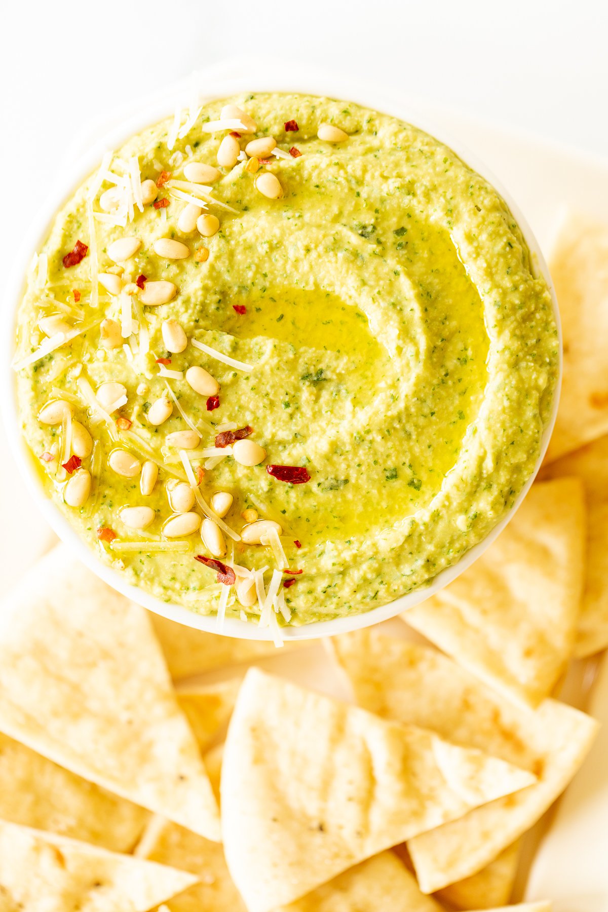 A white bowl full of basil pesto hummus, topped with pine nuts and parmesan.