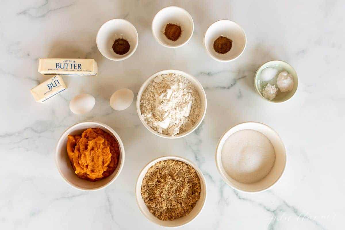 A marble surface with small white bowls filled with pumpkin bread recipe ingredients.