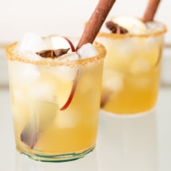 Clear glasses with apple cider margaritas, rimmed in sugar and garnished with a cinnamon stick on a marble surface.