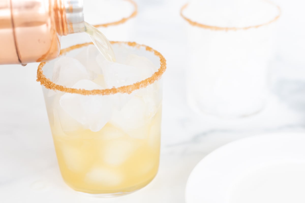 A shaker pouring an apple cider margarita into a cocktail glass.