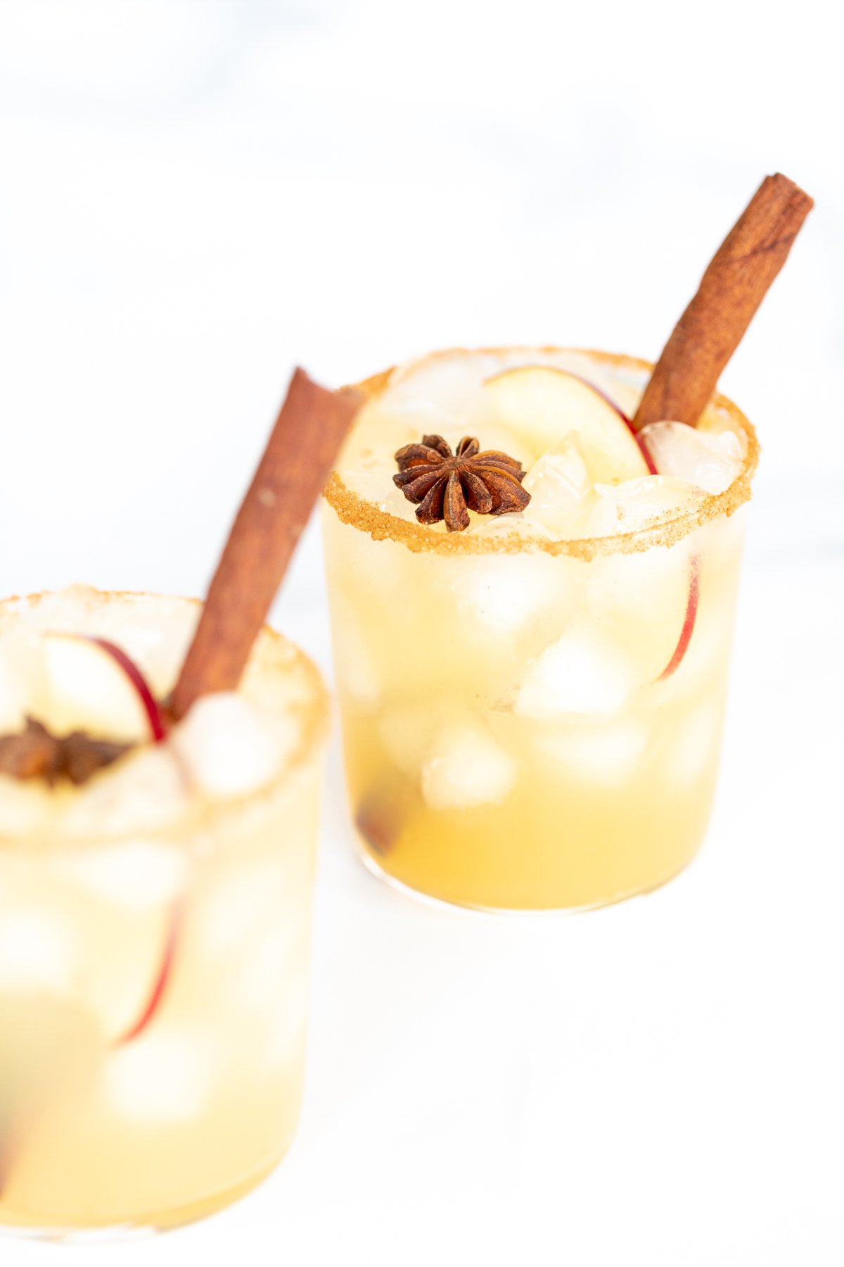 Clear glasses with apple cider margaritas, rimmed in sugar and garnished with a cinnamon stick on a marble surface.